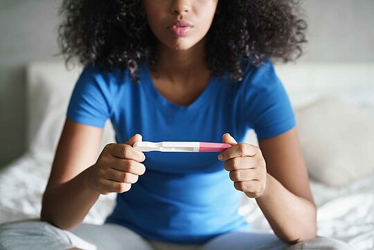 Frau sitzt auf Bett und hält einen Schwangerschaftstest in den Händen 