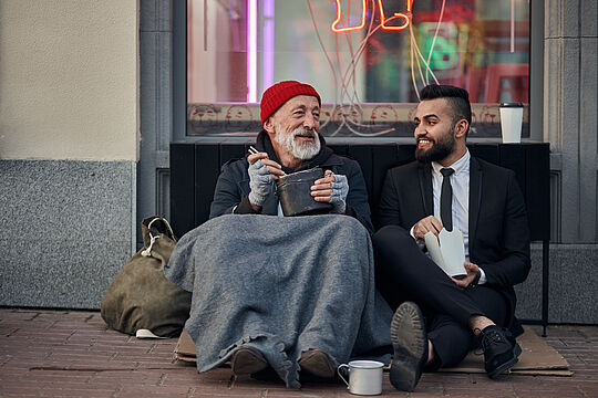 Wohnungsloser Mensch sitzt auf der Straße, neben ihm sitzt ein Mann im Anzug, beide lachen