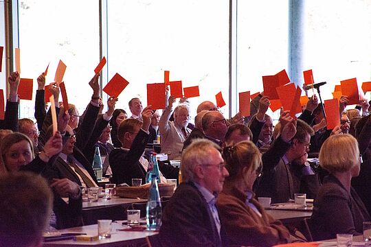 Blick in die Mitgliederversammlung der Diakonie Hessen. Die teilnehmenden Personen halten rote Abstimmungskarten in die Höhe.